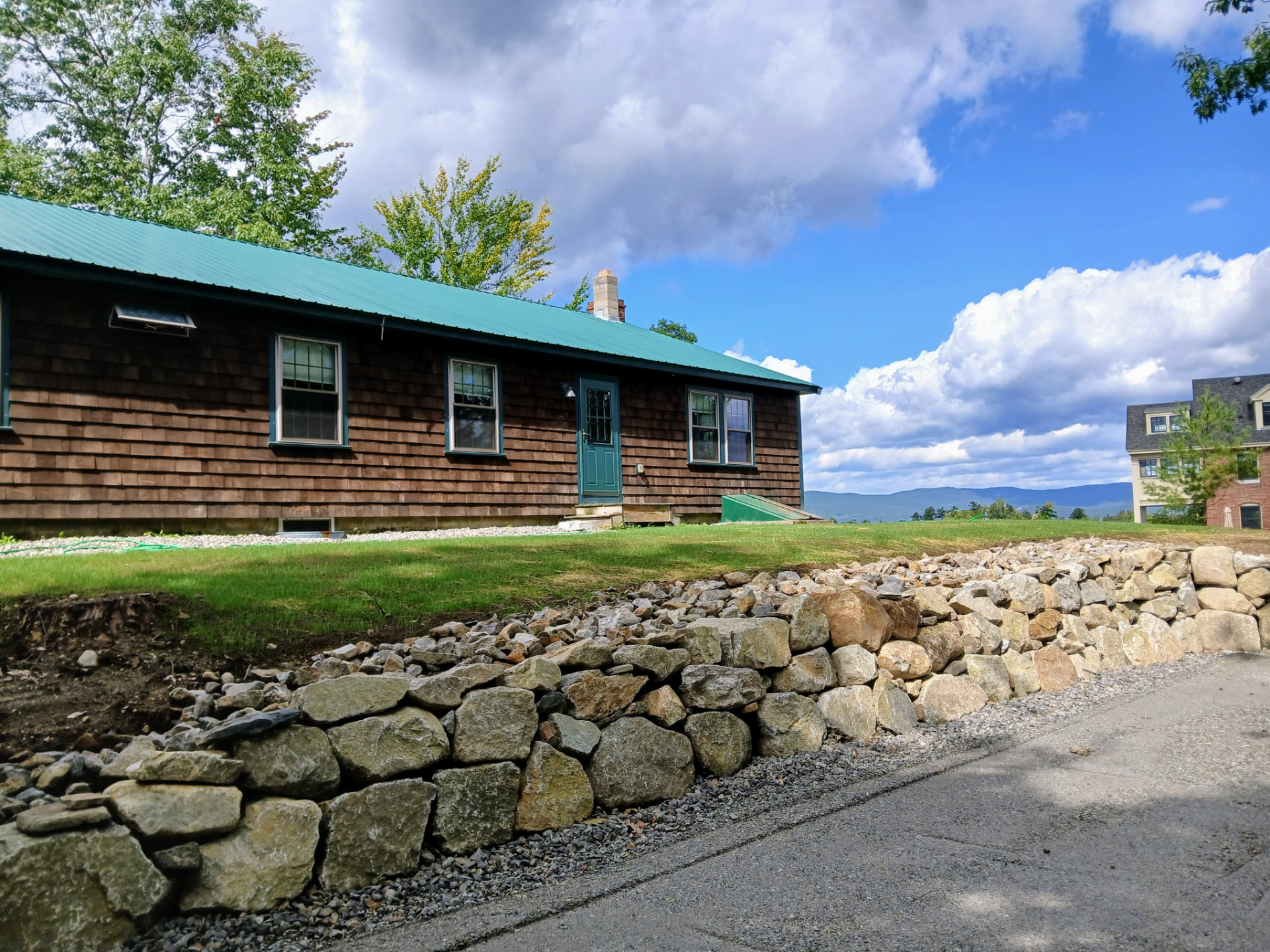 A single-story wood-shingled building with a green metal roof, standing above a stacked stone wall with hills beyond