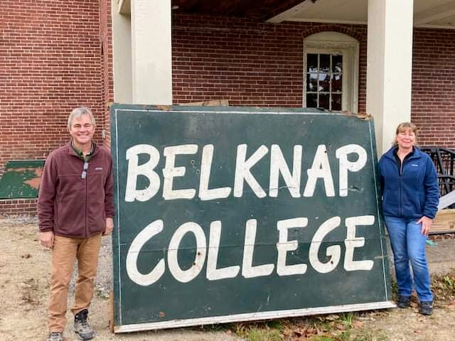Two people standing either side of the hand-painted Belknap College sign in front of the brick building